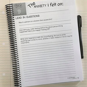 May include: A white notebook with a black spiral binding. The notebook has a black and white illustration of a brain on the cover. The text "The Anxiety I felt on:" is written in black at the top of the page. The page has a section titled "Lead In Questions:" with two questions. The first question is "Was it a person or a situation that caused this?" The second question is "How were you really feeling before the above person/thing happened? Were you happy? Feeling Off?" The page also has a section titled "Write your experience with this issue/feeling: Be sure to write anything that comes to mind, whether it feels related or not (it may be important!)" with lined space for writing. The bottom of the page has the word "CONTINUED" written in black with an arrow pointing to the right.