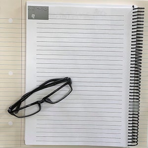 May include: A black framed pair of eyeglasses on a white lined notebook with a black spiral binding.