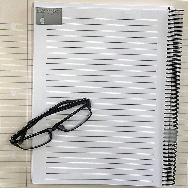 May include: A black framed pair of eyeglasses on a white lined notebook with a black spiral binding.