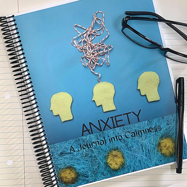May include: A blue journal with the title "Anxiety: A Journal into Calmness" in black text. The cover features three yellow felt heads with a tangled string of red and white yarn connected to the top head.