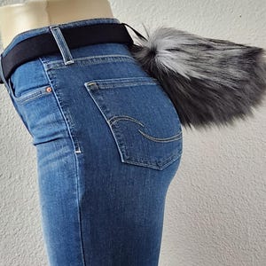 May include: A faux fur tail in shades of gray and white attached to a black belt. The belt is worn with blue denim jeans. The tail is fluffy and has a tapered shape, adding a playful accessory to the outfit.