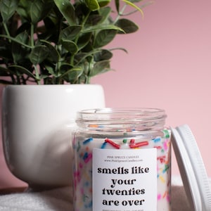 May include: A white candle jar with a white label that reads "smells like your twenties are over". The candle is decorated with colorful sprinkles and is hand-poured with soy wax.
