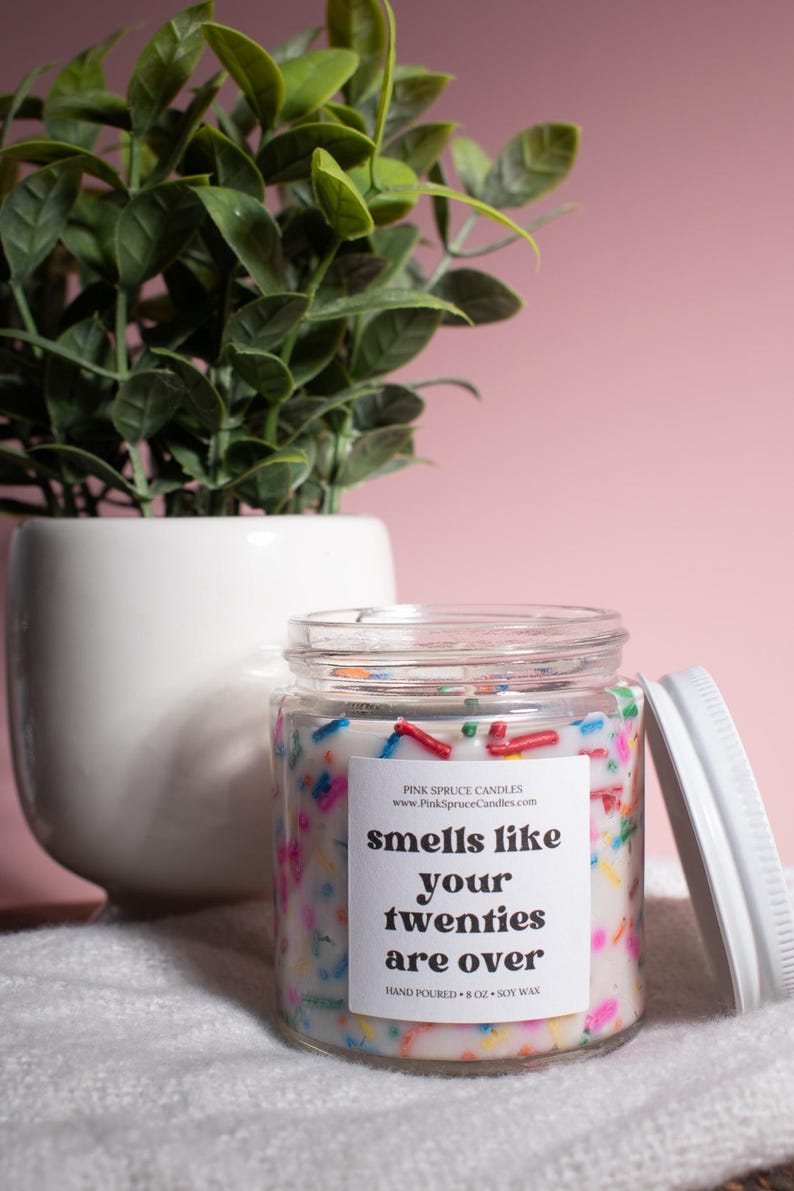 May include: A white candle jar with a white label that reads "smells like your twenties are over". The candle is decorated with colorful sprinkles and is hand-poured with soy wax.