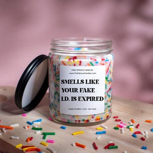 May include: A white soy candle in a clear glass jar with a black lid. The candle is decorated with colorful sprinkles and has a white label with the text "SMELLS LIKE YOUR FAKE I.D. IS EXPIRED" and "HAND POURED - 8 OZ - SOY WAX".