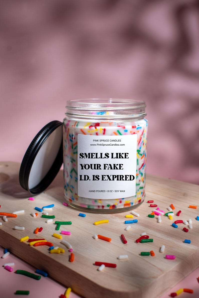 May include: A white soy candle in a clear glass jar with a black lid. The candle is decorated with colorful sprinkles and has a white label with the text "SMELLS LIKE YOUR FAKE I.D. IS EXPIRED" and "HAND POURED - 8 OZ - SOY WAX".