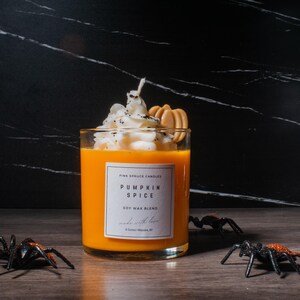 May include: A glass jar candle with a pumpkin spice scent. The candle is topped with whipped cream and a graham cracker. The label reads "Pink Spruce Candles Pumpkin Spice Soy Wax Blend Made with Love".