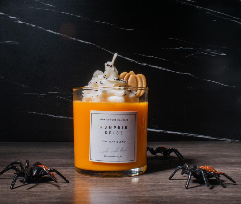 May include: A glass jar candle with a pumpkin spice scent. The candle is topped with whipped cream and a graham cracker. The label reads "Pink Spruce Candles Pumpkin Spice Soy Wax Blend Made with Love".