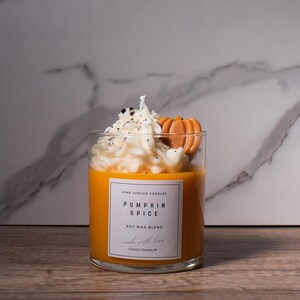 May include: A glass jar candle with a white soy wax blend topped with a whipped cream-like wax and a small pumpkin-shaped wax accent. The label reads "Pink Spruce Candles Pumpkin Spice Soy Wax Blend made with love 8 Ounces | Louisville, KY"