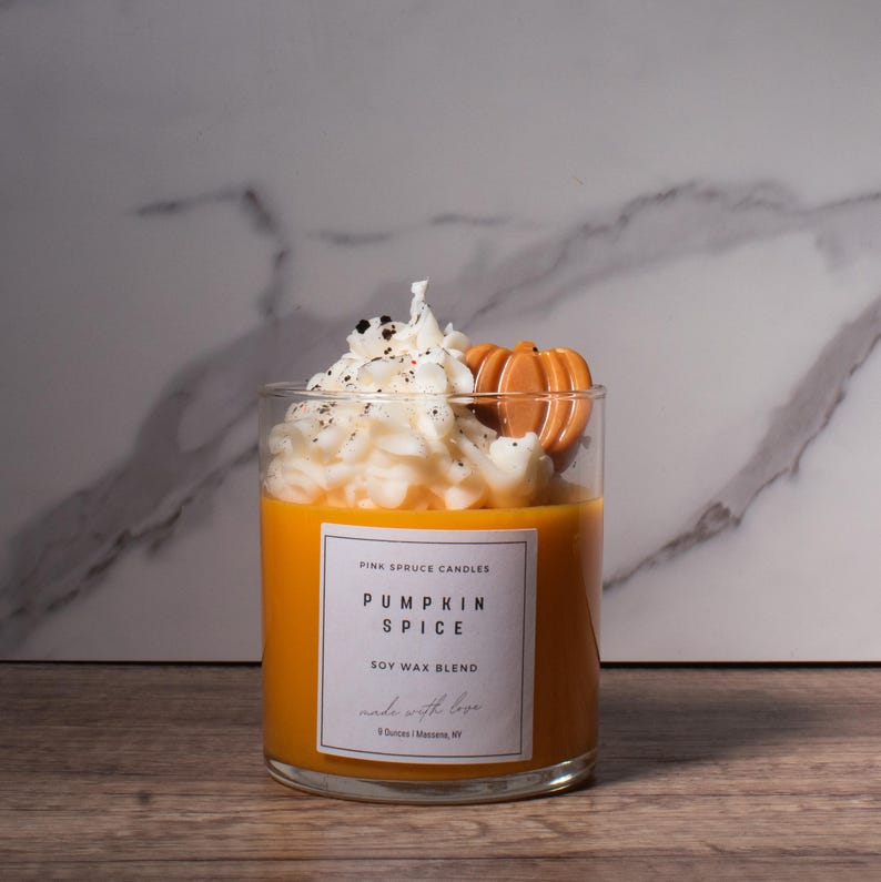 May include: A glass jar candle with a white soy wax blend topped with a whipped cream-like wax and a small pumpkin-shaped wax accent. The label reads "Pink Spruce Candles Pumpkin Spice Soy Wax Blend made with love 8 Ounces | Louisville, KY"
