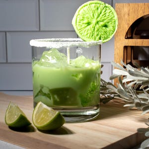 May include: A green cocktail with ice in a glass rimmed with sugar. A lime wedge is on top of the glass. Two lime wedges are on a wooden cutting board.