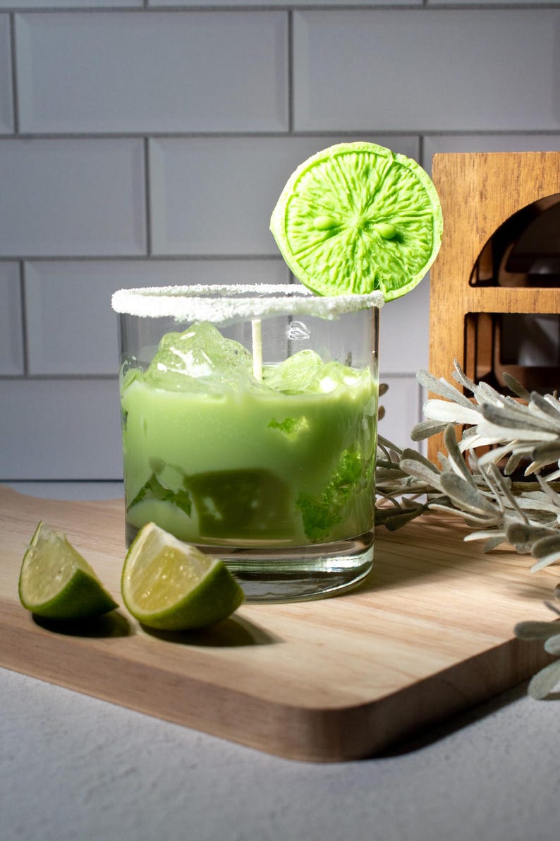 May include: A green cocktail with ice in a glass rimmed with sugar. A lime wedge is on top of the glass. Two lime wedges are on a wooden cutting board.