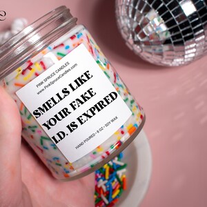 May include: A white candle with a colorful sprinkle design and a label that reads "Smells Like Your Fake I.D. Is Expired". The candle is hand-poured and made with soy wax. It is 8 ounces.