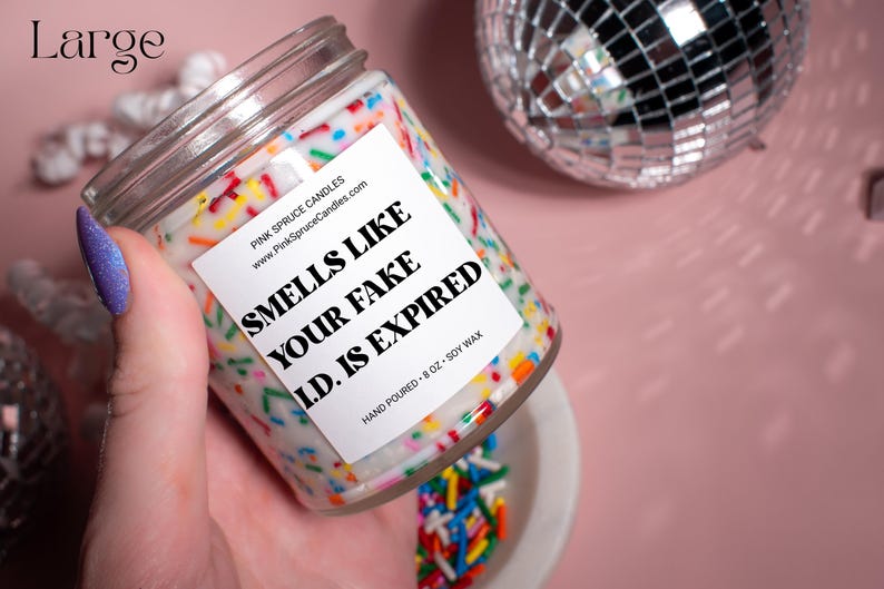 May include: A white candle with a colorful sprinkle design and a label that reads "Smells Like Your Fake I.D. Is Expired". The candle is hand-poured and made with soy wax. It is 8 ounces.