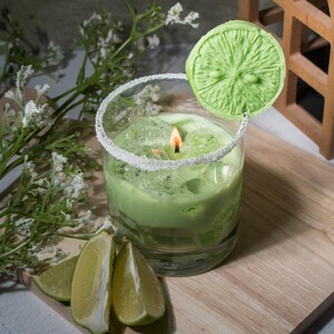 May include: A green candle in a glass jar with a sugared rim, topped with a green lime slice. The candle is surrounded by lime wedges on a wooden surface.
