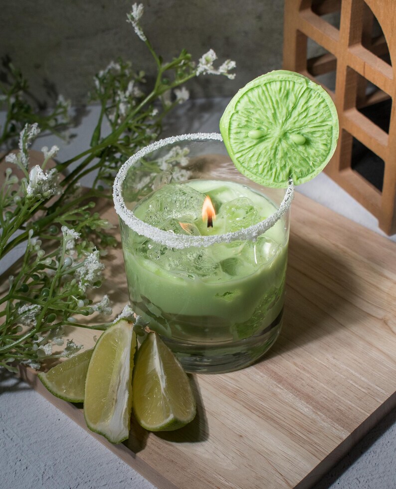 May include: A green candle in a glass jar with a sugared rim, topped with a green lime slice. The candle is surrounded by lime wedges on a wooden surface.