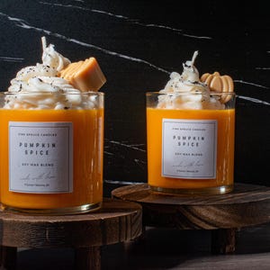 May include: Two pumpkin spice scented soy wax candles in glass jars with a white whipped cream topping and a pumpkin spice topping. The candles are on wooden stands.