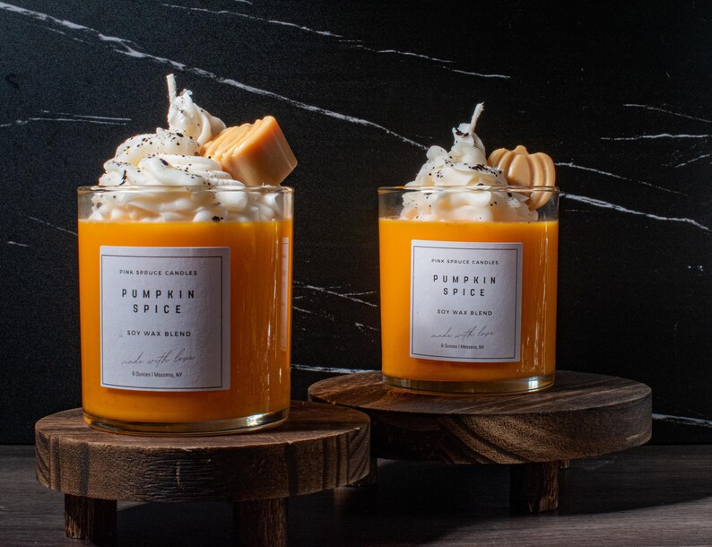 May include: Two pumpkin spice scented soy wax candles in glass jars with a white whipped cream topping and a pumpkin spice topping. The candles are on wooden stands.