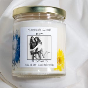 Może przedstawiać: Świeca z białego wosku sojowego w przezroczystym szklanym słoiku ze złotą pokrywką. Etykieta zawiera napis Pink Spruce Candles, Ruby, Bridesmaid, Soy 8 oz Cake Scented oraz czarno-białe zdjęcie. Widoczne są prasowane niebieskie i ż&oacute;łte kwiaty.