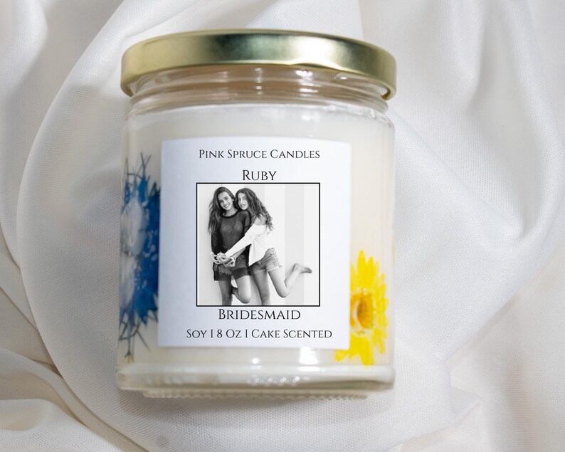 Może przedstawiać: Świeca z białego wosku sojowego w przezroczystym szklanym słoiku ze złotą pokrywką. Etykieta zawiera napis Pink Spruce Candles, Ruby, Bridesmaid, Soy 8 oz Cake Scented oraz czarno-białe zdjęcie. Widoczne są prasowane niebieskie i ż&oacute;łte kwiaty.