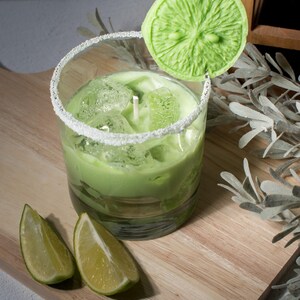 May include: A green candle in a glass jar with a sugared rim, topped with a green lime slice candle. Two lime slices are on a wooden surface.