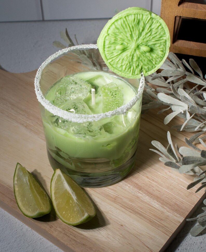 May include: A green candle in a glass jar with a sugared rim, topped with a green lime slice candle. Two lime slices are on a wooden surface.