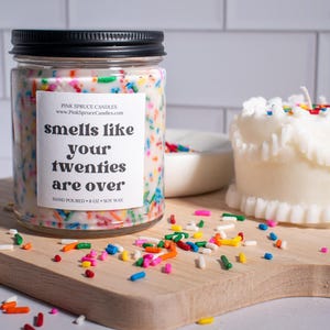May include: A white candle with colorful sprinkles on top in a glass jar with a black lid. The label on the jar reads "smells like your twenties are over". The candle is sitting on a wooden cutting board with white sprinkles scattered around it.
