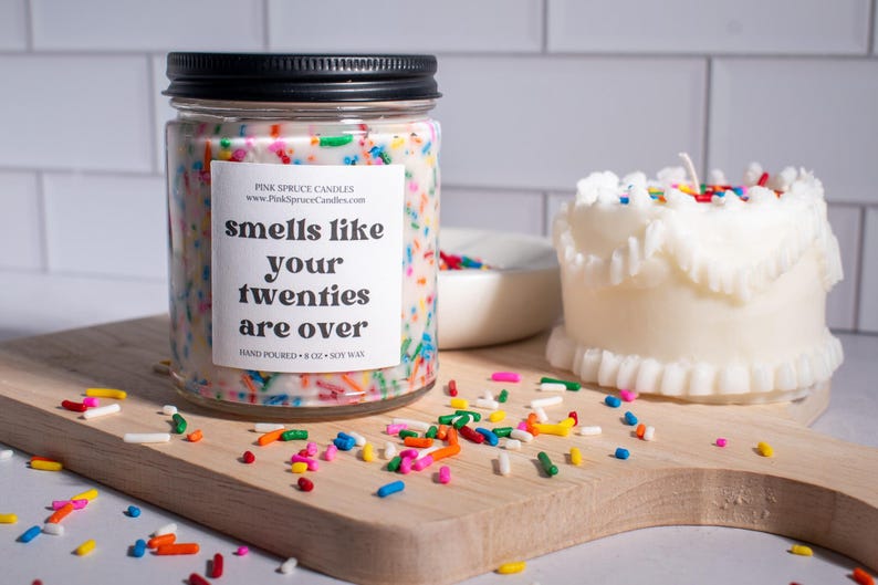 May include: A white candle with colorful sprinkles on top in a glass jar with a black lid. The label on the jar reads "smells like your twenties are over". The candle is sitting on a wooden cutting board with white sprinkles scattered around it.