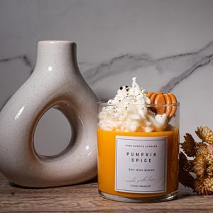 May include: A glass candle jar with a soy wax blend. The candle is topped with a white whipped cream-like wax and a small orange pumpkin-shaped wax decoration. The label reads "Pumpkin Spice Soy Wax Blend" and "Pink Spruce Candles".