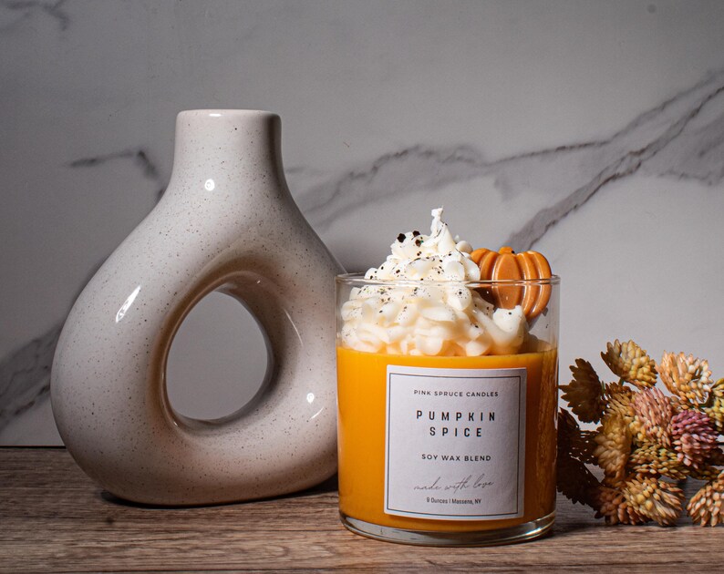 May include: A glass candle jar with a soy wax blend. The candle is topped with a white whipped cream-like wax and a small orange pumpkin-shaped wax decoration. The label reads "Pumpkin Spice Soy Wax Blend" and "Pink Spruce Candles".