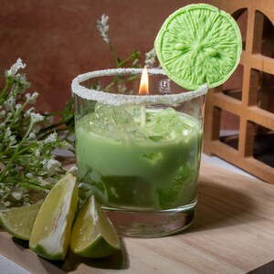 May include: A green candle in a glass jar with a white rim and a lime slice on top. The candle is lit and the wax is melted. There are lime wedges on a wooden cutting board in the foreground.