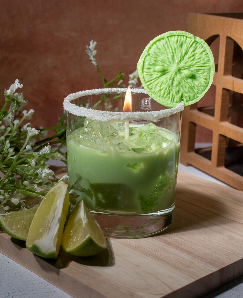 May include: A green candle in a glass jar with a white rim and a lime slice on top. The candle is lit and the wax is melted. There are lime wedges on a wooden cutting board in the foreground.