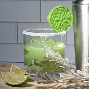 May include: A green lime wedge garnish sits on the rim of a glass filled with a green liquid and ice. The glass is on a wooden cutting board with a sprig of greenery and lime wedges.