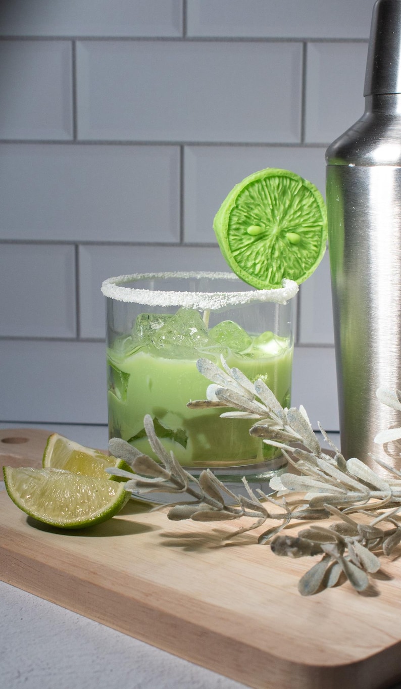 May include: A green lime wedge garnish sits on the rim of a glass filled with a green liquid and ice. The glass is on a wooden cutting board with a sprig of greenery and lime wedges.