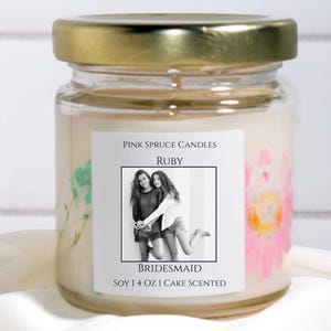 Może przedstawiać: Przezroczysty szklany słoik na świecę ze złotą pokrywką, wypełniony kremowym woskiem sojowym. Etykieta zawiera napis Pink Spruce Candles, Ruby, Bridesmaid oraz Soy 4 oz Cake Scented, czarno-białe zdjęcie i prasowane kwiaty.
