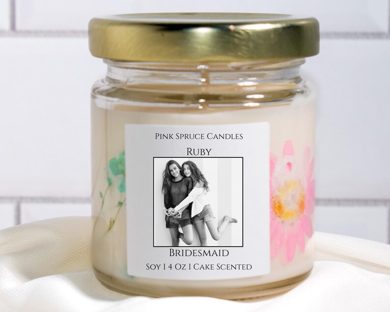 Może przedstawiać: Przezroczysty szklany słoik na świecę ze złotą pokrywką, wypełniony kremowym woskiem sojowym. Etykieta zawiera napis Pink Spruce Candles, Ruby, Bridesmaid oraz Soy 4 oz Cake Scented, czarno-białe zdjęcie i prasowane kwiaty.