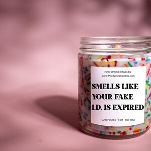 May include: A white candle jar with a colorful sprinkle design and a white label that reads "SMELLS LIKE YOUR FAKE I.D. IS EXPIRED". The jar is 8 oz and made with soy wax.