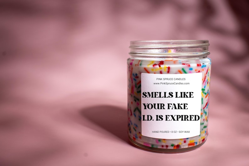 May include: A white candle jar with a colorful sprinkle design and a white label that reads "SMELLS LIKE YOUR FAKE I.D. IS EXPIRED". The jar is 8 oz and made with soy wax.