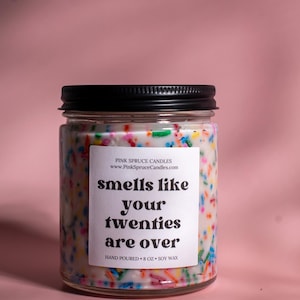 May include: A glass candle jar with a black lid and a white label that says "smells like your twenties are over". The candle is filled with white wax and colorful sprinkles. The jar is 8 ounces and made with soy wax.