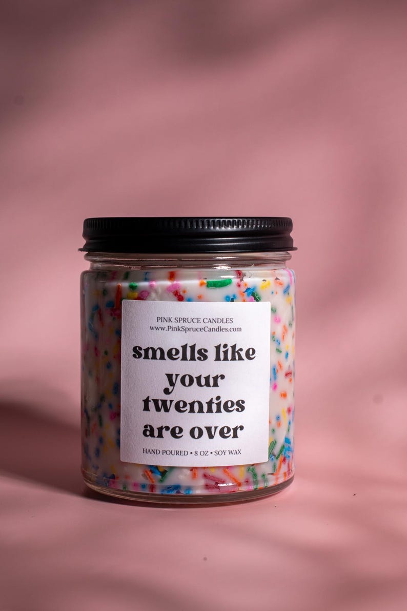 May include: A glass candle jar with a black lid and a white label that says "smells like your twenties are over". The candle is filled with white wax and colorful sprinkles. The jar is 8 ounces and made with soy wax.