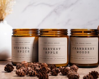 May include: Three amber glass candle jars with brown paper labels. The labels read "Strudel + Spice", "Harvest Apple", and "Cranberry Woods". The candles are surrounded by pine cones on a wooden surface.