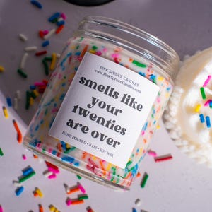 May include: A white candle with colorful sprinkles in a glass jar. The label reads "smells like your twenties are over" and "Pink Spruce Candles".
