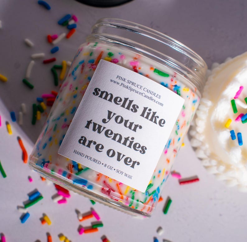 May include: A white candle with colorful sprinkles in a glass jar. The label reads "smells like your twenties are over" and "Pink Spruce Candles".