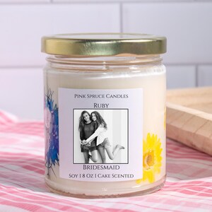 Może przedstawiać: Przezroczysty szklany słoik na świecę ze złotą pokrywką i białym woskiem. Etykieta zawiera napis Pink Spruce Candles, Ruby, Bridesmaid, czarno-białe zdjęcie dw&oacute;ch os&oacute;b oraz tekst Soy 8 oz Cake Scented i grafikę ż&oacute;łtych kwiat&oacute;w.