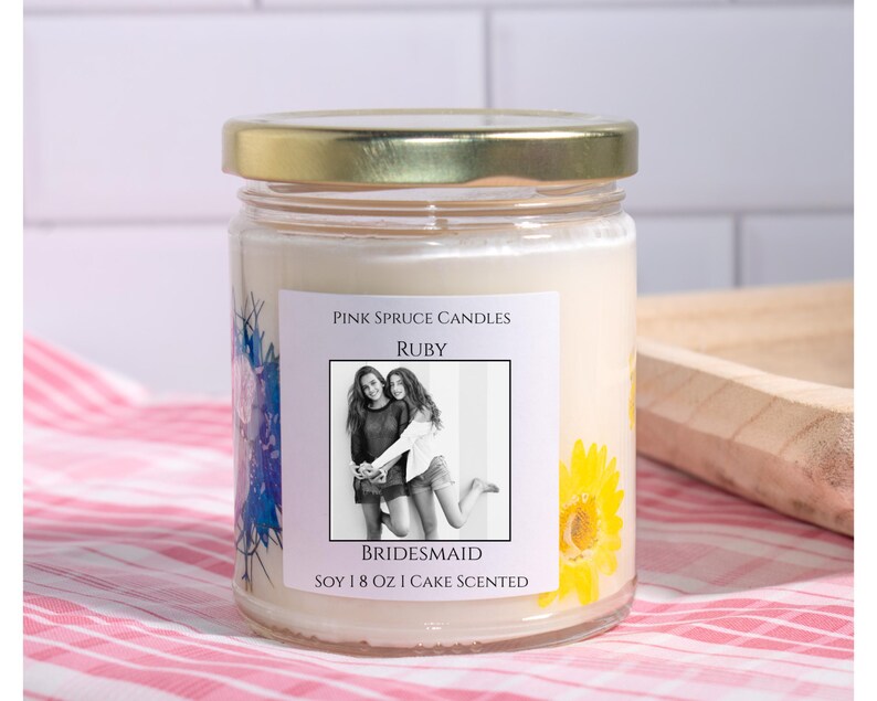 Może przedstawiać: Przezroczysty szklany słoik na świecę ze złotą pokrywką i białym woskiem. Etykieta zawiera napis Pink Spruce Candles, Ruby, Bridesmaid, czarno-białe zdjęcie dw&oacute;ch os&oacute;b oraz tekst Soy 8 oz Cake Scented i grafikę ż&oacute;łtych kwiat&oacute;w.