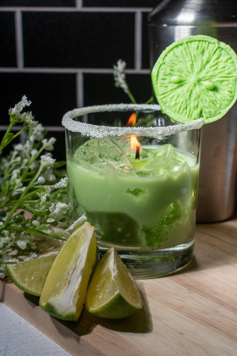 May include: A green candle in a glass jar with a white rim and a lime wedge on top. The candle is surrounded by white flowers and lime slices.