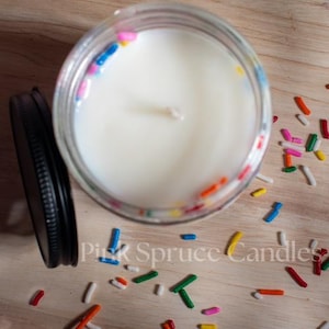 May include: A white candle with colorful sprinkles embedded in the wax. The candle is in a clear glass jar with a black lid. The jar is surrounded by more sprinkles on a wooden surface.