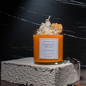 May include: A glass jar candle with a pumpkin spice scent. The candle is topped with a white whipped cream-like topping and a small orange cookie. The jar has a white label with the text "Pink Spruce Candles Pumpkin Spice Soy Wax Blend Made with Love"