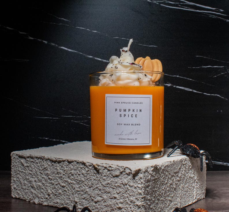 May include: A glass jar candle with a pumpkin spice scent. The candle is topped with a white whipped cream-like topping and a small orange cookie. The jar has a white label with the text "Pink Spruce Candles Pumpkin Spice Soy Wax Blend Made with Love"