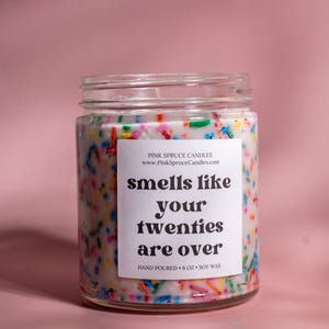 May include: A glass jar candle with a white label that reads "smells like your twenties are over". The candle is decorated with colorful sprinkles and has a white background. The candle is hand-poured and made with soy wax.