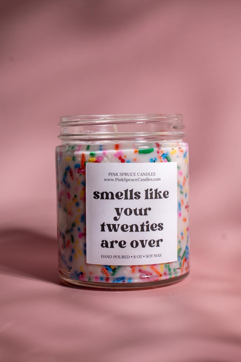 May include: A glass jar candle with a white label that reads "smells like your twenties are over". The candle is decorated with colorful sprinkles and has a white background. The candle is hand-poured and made with soy wax.